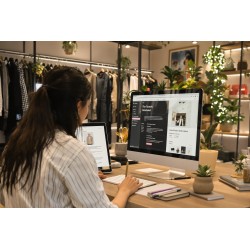 Personne assise à un bureau dans une boutique, travaillant sur ordinateur et tablette avec code et texte, entourée de racks de v