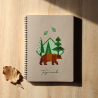 Carnet de notes brun à spirale posé sur planche en bois, couverture illustrée d’un ours et d’éléments forestiers verts.