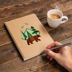 Carnet de notes en kraft avec logo d’ours brun et forêt géométrique, stylo à la main près d’une tasse de café.